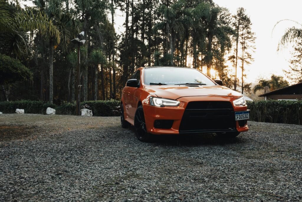 An orange Mitsubishi Lancer parked on a gravel road with a forest backdrop during dusk.